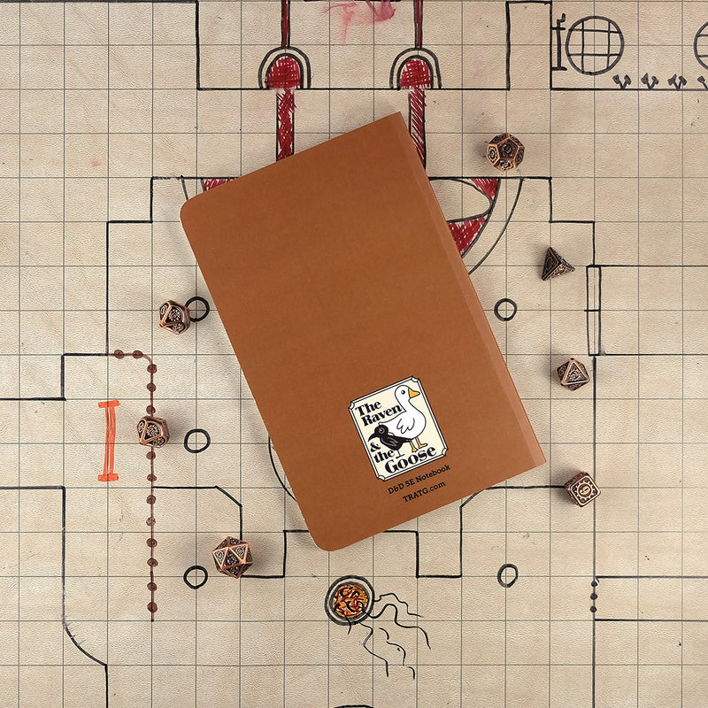 The white back cover of the artificer character notebook with "The Raven and the Goose" logo on the bottom. The notebook sits on a grid map surrounded by dice.