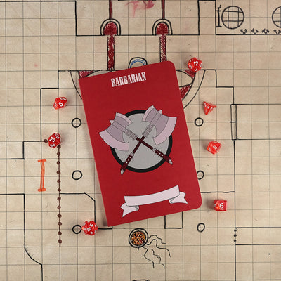 The front cover of a large barbarian notebook with a bright red cover and an illustration of two axes crossing one another. The notebook sits on a grid map surrounded by dice.