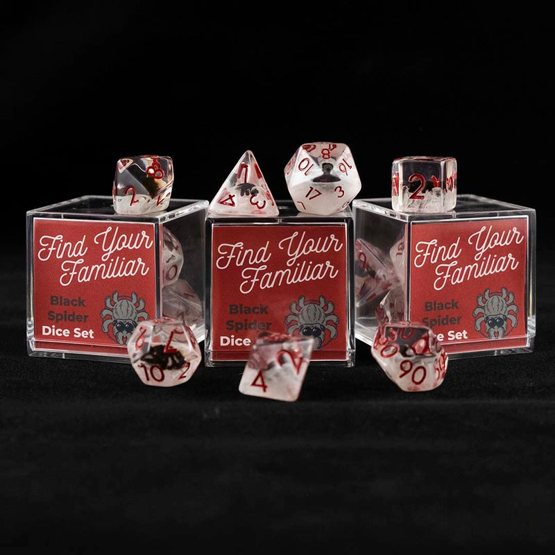 A set of clear and cloudy polyhedral dice with black spider inclusions sitting on top and around a three small acrylic boxes with the words "Find Your Familiar" and "Black Spider Dice Set" on a black background.