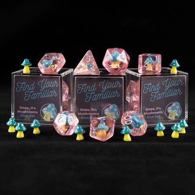 A set of pink polyhedral dice with blue and pink cap mushroom inclusions sitting on top and around a three small acrylic boxes with the words "Find Your Familiar" and "Oops, It's Mushrooms Dice Set" with small mushrooms on a black background.
