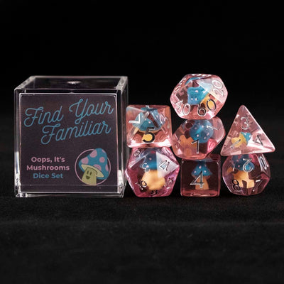 A set of pink polyhedral dice with blue and pink cap mushroom inclusions sitting to the right of a small acrylic box with the words "Find Your Familiar" and "Oops, It's Mushrooms Dice Set" on a black background.