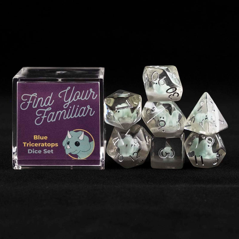 A set of clear polyhedral dice with blue triceratops inclusions sitting to the right of a small acrylic box with the words "Find Your Familiar" and "Blue Triceratops Dice Set" on a black background.