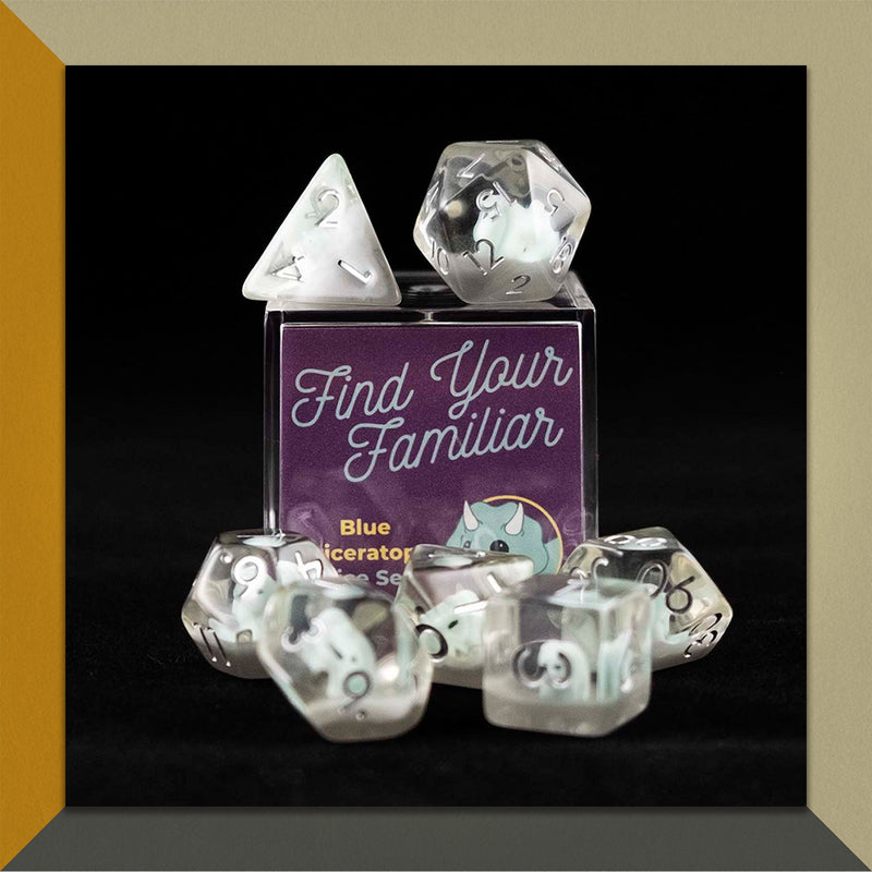 A set of clear polyhedral dice with blue triceratops inclusions sitting on top and around a small acrylic box with the words "Find Your Familiar" and "Blue Triceratops Dice Set" on an orange, cream and grey background.