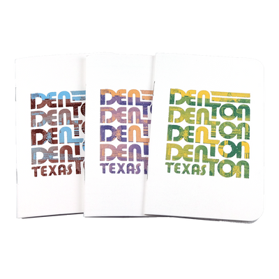 The front covers of three Denton notebooks with the word "Denton" stacked four times and the word "Texas" underneath in maroon and blue, purple and orange, and green and yellow duotones. The notebooks are on a transparent background.