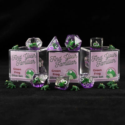 A set of clear and purple polyhedral dice with green frog inclusions sitting to the right of a small acrylic box with the words "Find Your Familiar" and "Green Frog Dice Set" on a black background.