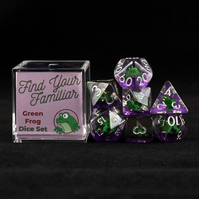 A set of clear and purple polyhedral dice with green frog inclusions sitting to the right of a small acrylic box with the words "Find Your Familiar" and "Green Frog Dice Set" on a black background.