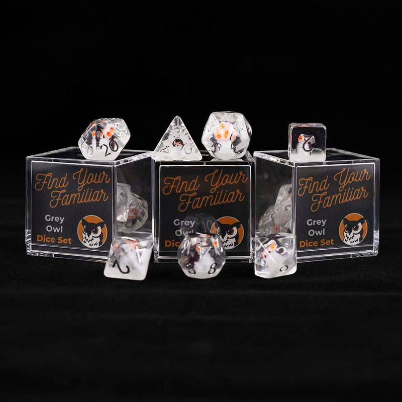 A set of clear polyhedral dice with grey owl inclusions sitting on top and around a three small acrylic boxes with the words "Find Your Familiar" and "Grey Owl Dice Set" on a black background.