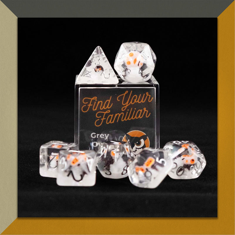 A set of clear polyhedral dice with grey owl inclusions sitting on top and around a small acrylic box with the words "Find Your Familiar" and "Grey Owl Dice Set" on an orange, cream and grey background.