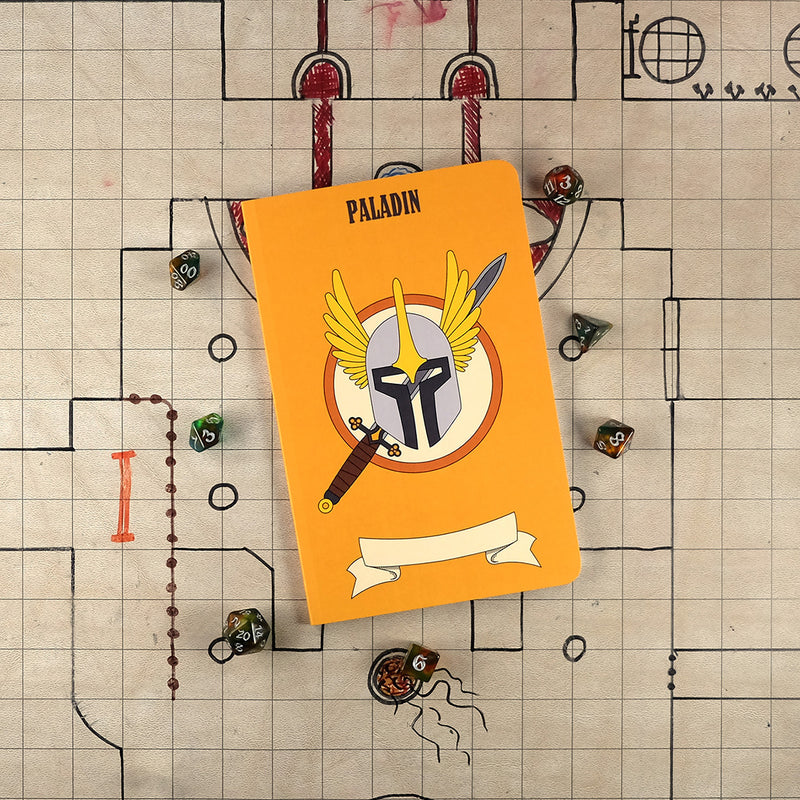 The front cover of a large paladin notebook with an orange cover and an illustration of sword and a winged helm . The notebook sits on a grid map surrounded by dice.