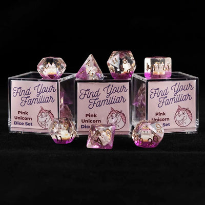 A set of clear and purple polyhedral dice with pink unicorn inclusions sitting on top and around a three small acrylic boxes with the words "Find Your Familiar" and "Pink Unicorn Dice Set" on a black background.