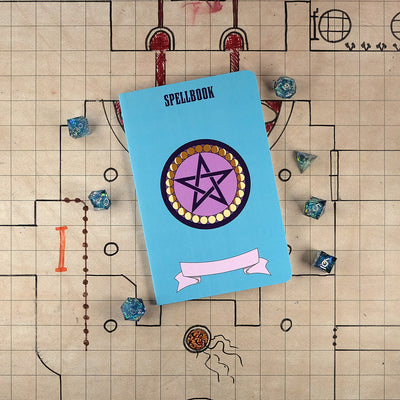 The front cover of a large spellbook notebook with a bright blue cover and an illustration of a purple pentacle surrounded by yellow circles with shadows for the different phases of the moon. The notebook sits on a grid map surrounded by dice.