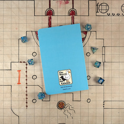 The bright blue back cover of the spellbook notebook with "The Raven and the Goose" logo on the bottom. The notebook sits on a grid map surrounded by dice.