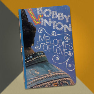 Photo of a 5.25” x 8.25” sketchbook made from an upcycled Bobby Vinton “Melodies of Love” album cover on an orange, cream and grey background.